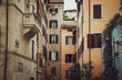 © Hanne - A narrow street lined with tall, closely packed buildings in warm tones. The buildings have classical architecture with windows, shutters, and balconies. Greenery is visible, and the sky is partially