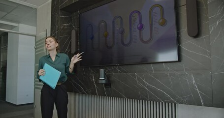 Wall Mural - A young businesswoman using a pen to point at a graph displayed on a monitor. She is holding a light blue folder and is wearing a green shirt and black pants.