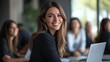 © Haram - Confident businesswoman in professional attire, smiling at her desk with a laptop while team members collaborate in a modern office with bright lighting, large windows, and a productive atmosphere.