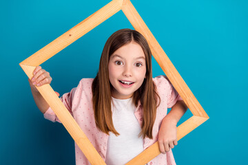 Wall Mural - Smiling girl holding a wooden frame against a bright blue background, expressing joy and creativity