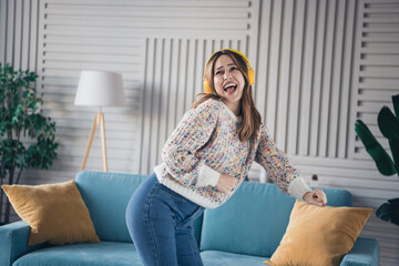 Wall Mural - Joyful young woman enjoying music and dancing energetically in a cozy living room with a colorful sweater and headphones on