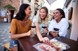 © CarlosBarquero - Three cheerful female friends are laughing and enjoying a glass of red wine with a charcuterie board, spending quality time together in a picturesque Italian town