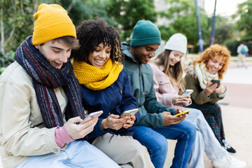 Wall Mural - Young group of diverse teen friends using mobile phones sitting on bench. Multiracial Gen Z student people in warm clothes browsing social media and connecting online. Youth and technology concept