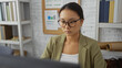 © Krakenimages.com - Young chinese woman working at an office desk with focus, wearing glasses and a green blazer, with shelves and a bulletin board in the background, creating a professional and modern workspace.