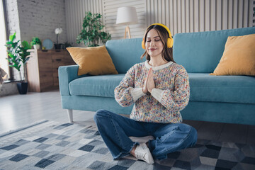 Wall Mural - Young woman meditating in a cozy apartment, wearing headphones, practicing mindfulness in the living room