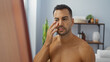 © Krakenimages.com - Handsome hispanic man with a beard examining his face in a spa room's wellness center, reflecting an indoor beauty and wellness ambiance