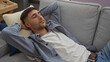 © Krakenimages.com - Young man relaxing on a sofa indoors with his arms behind his head, sporting casual attire, conveying a sense of comfort and tranquility in a modern home setting.