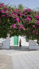 Wall Mural - Woman standing under vibrant bougainvillea in haria, lanzarote, dressed in black with a white bag, next to a white building with a green door, capturing the essence of outdoor beauty.