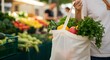© youns - Un primer plano de la mano de una persona sosteniendo una bolsa de compras reutilizable con verduras al fondo en un mercado de agricultores local