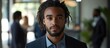 © StockKing - Professional young man with dreadlocks in a dark suit standing in a well-lit modern office space, with blurred colleagues in the background.