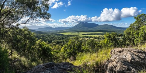 Aufkleber - Lush green valley with distant mountains under a blue sky dotted with fluffy clouds, rocky foreground providing depth to the serene landscape view.