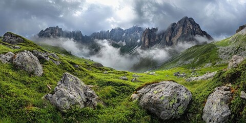 Aufkleber - Panoramic view of majestic mountains shrouded in mist under a dramatic cloudy sky, with lush green hills and large rocky foreground elements.
