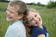 © Pujan - Joyful children outdoors, laughing expressions, back-to-back pose, grassy field background, natural light, candid moment, childhood happiness, blue and white clothing, summer day