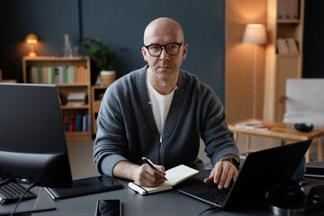 Portrait of middle aged man with shaved head looking at camera with calm expression on face, while writing in notebook working online on laptop in office