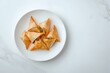 © Yuliia - Golden crispy phyllo triangles filled with savory ingredients served on white plate against marble background, overhead view of Mediterranean appetizers.