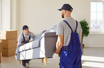 Male movers team of moving service carrying heavy sofa together shipping furniture in new apartment. Men workers from delivery service placing modern couch in new house. Moving day concept.