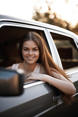 Wall Mural - Young girl smiling from car window in sunny setting, showcasing joy, happiness, and a carefree attitude with warm color tones and natural background.