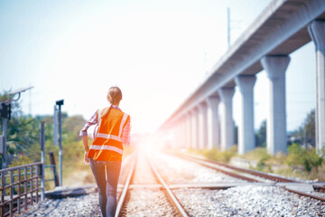 Wall Mural - White female Rail transportation engineer in safety vest walking back along the railway track with the light and moterway background.