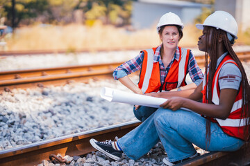 Wall Mural - Black and White female Rail transportation engineers in safety vest and hardhat check the construction structure of the railway track while holding blueprint