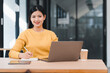© kenchiro168 - Woman in yellow sweater working on laptop while taking notes, sitting at wooden desk with coffee cup in modern office.