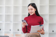 © kenchiro168 - Young Asian woman in red sweater smiles while using her smartphone and holding documents in modern office.