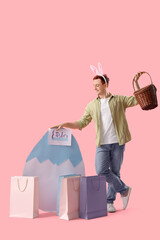 Wall Mural - Young man in bunny ears with basket, paper egg and shopping bags on pink background. Easter Sale