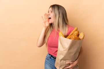 Poster - Young blonde woman holding a bag full of breads isolated on beige background shouting with mouth wide open