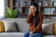 © Liubomir - Asian woman experiencing ear pain on couch holding her ear with discomfort. Casual wear at home conveying discomfort, health issues, or earache pain. Indoor domestic setting.