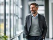 © tao - Handsome smiling businessman in business casual stands in office corridor, laughing at camera with brown hair and light stubble beard. Modern glass wall open space in background.