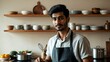© Arceli - A young South Asian man cooking in an apron in a home kitchen in closeup portrait on a plain light beige background