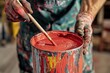 © whitestorm - Painter stirring a bucket of red paint with a stick, wearing paint-splattered gloves and clothing in a creative studio environment, getting ready to create art.