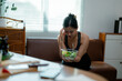 © PaeGAG - Young asian woman wincing from headache, gripping salad bowl while resting on home couch, depicting health struggles and nutrition challenges