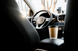 © ShvedKristina - A recyclable coffee cup and lid are on the armrest next to the driver in the car interior, view from the back seat of the car. Coffee in the car. Sun rays in the car. Traveling in the car with coffee