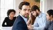 © fizkes - Company boss close up portrait. Profile picture of young handsome businessman, client or investor posing seated at desk, businesspeople on background, take part in formal meeting or negotiations event