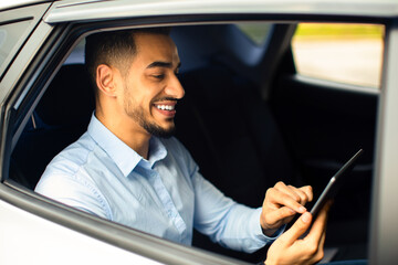 Wall Mural - Cheerful middle eastern businessman checking emails while going to airport by taxi, sitting on car back seat and using digital tablet, closeup photo, shot from outside, side view, copy space