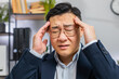 © Andrii Iemelianenko - Close-up face of tired ill Asian businessman suffering from headache problem tension and migraine, stress at office desk. Mature freelancer broker worker man works on laptop notebook. Overworking