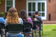 © evgenia_lo - Several people seated in chairs outdoors, attentively listening to a speaker at a gathering taking place in a serene environment