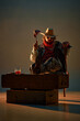© master1305 - Cowboy sitting with foot on chest, holding raw meat on skewer, glass of whiskey on wooden trunk. Man with intense gaze, in hat, red bandana and denim jacket on dark background. American culture
