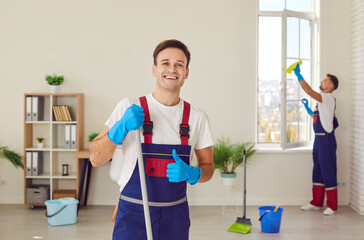 Wall Mural - Happy professional janitor. Two commercial cleanup service workers use cleaning tools and equipment, wash windows, tidy up living room. Cheerful smiling man satisfied with good salary shows thumbs up