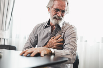 Senior man in spectacles presses hand to chest has heart attack suffers from unbearable chest pain closes eyes wears glasses sitting on the chair in living room. People age and problems with health.