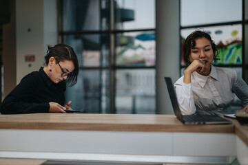 Wall Mural - Two professional women are engaged in work at a contemporary office setting, using laptops and smart phones. The image conveys a sense of focus and collaboration in a business environment.