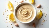Top view tahini sauce in bowl surrounded by sesame seeds, lemon, garlic on bright white surface. Creamy tasty dish, blended eastern culinary dressing for vegan, healthy lunch.