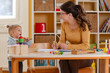 © lordn - A happy preschool teacher interacts with a cheerful toddler girl in a bright and organized classroom, fostering a positive learning environment