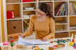 © lordn - A focused preschool teacher in glasses writes notes at a desk in a bright classroom, surrounded by educational toys and learning materials