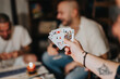 © qunica.com - Close-up of a hand holding playing cards, while a group of friends enjoys a card game in the background, fostering connection and relaxation.