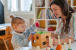 A young woman and a little girl play with educational toys at a preschool or daycare, engaging in learning activities with a shape sorter in a bright, cheerful classroom