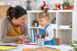 © lordn - A young girl in a denim dress holds a colored pencil while interacting with her smiling teacher in a bright classroom, engaging in a fun and educational drawing activity