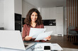 © Stock 4 You - Smiling latin hispanic young business woman working on laptop computer reading financial bills document at home office. Accountant entrepreneur manager businesswoman doing paperwork using pc remotely
