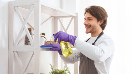 Domestic Cleaning. Young Man In Apron Cleans Dust From Shelf Indoors, Tidying Apartment, Copy Space