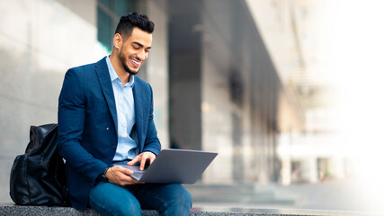 Positive middle eastern guy in suit with backpack sitting on the street, using laptop and smiling, young man entrepreneur checking presentation on computer before business meeting, copy space